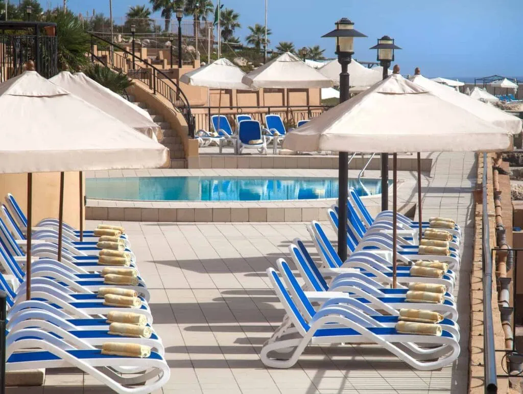 Discover how to beat the summer heat in Malta at our stunning poolside area. Relax on blue and white lounge chairs under crisp white umbrellas, with rolled yellow towels waiting for you. Take a dip in the small pool, surrounded by palm trees and offering a glimpse of the distant sea.