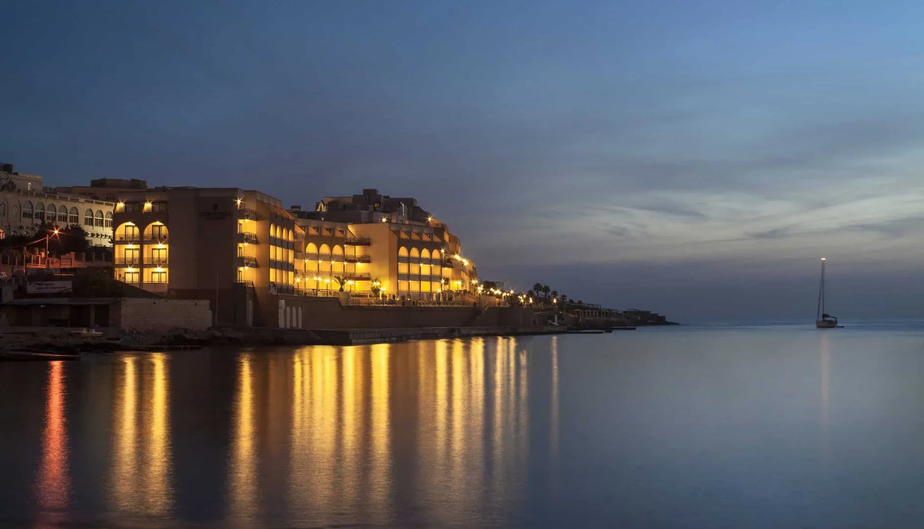 Hotel buildings with illuminated windows line a calm waterfront at dusk, reflecting on the water; a single boat floats in the distance under a cloudy sky.