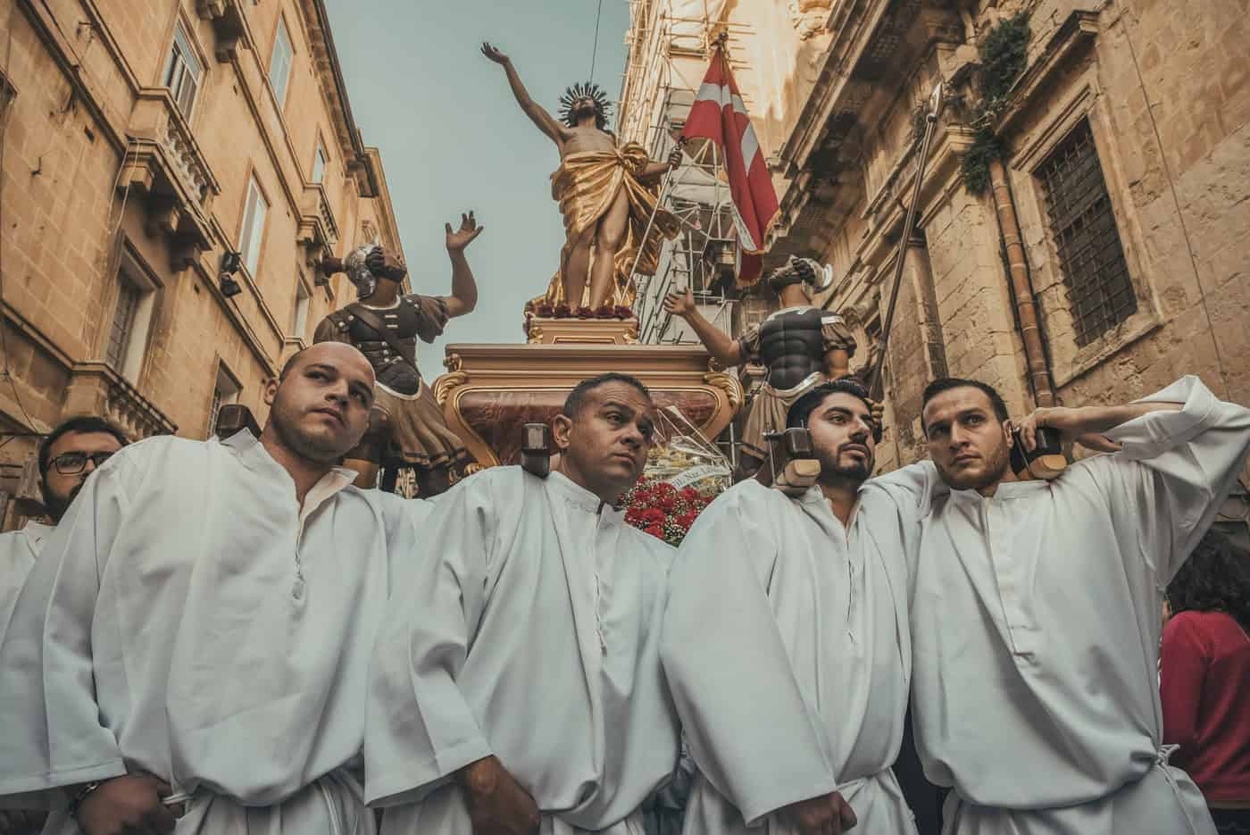 During Easter in Malta, four men in white robes carefully carry a religious statue through a narrow street. Buildings line the road, and onlookers are partially visible, capturing the essence of this traditional daytime procession.