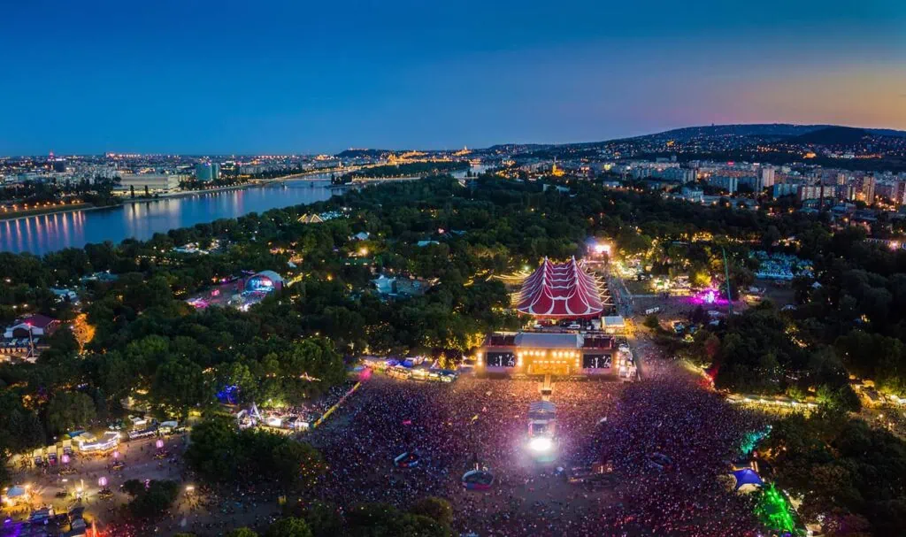 Aerial view of a large outdoor music festival at dusk, with a stage surrounded by a crowd, colorful lights, trees, a river, and a city skyline in the background.