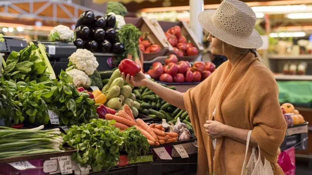 Amidst Malta's best farmers markets, a woman in a straw hat and brown shawl thoughtfully selects a red bell pepper. As she explores the vibrant stand, brimming with carrots, cucumbers, and leafy greens, she enjoys the bounty of fresh produce on offer.