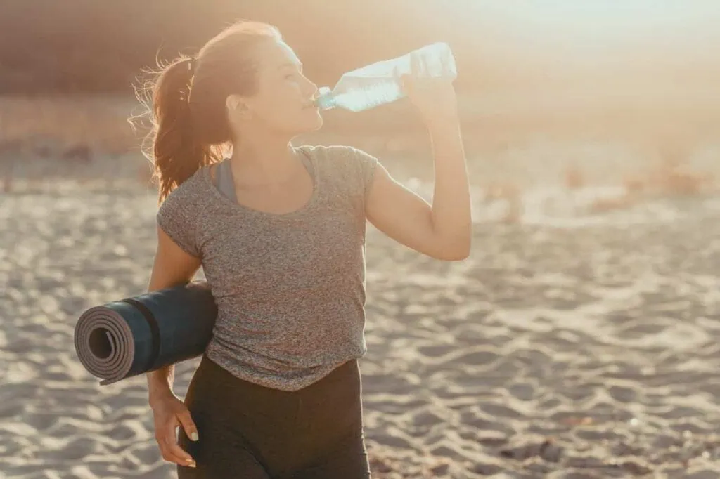 A woman stands on a sunny beach in Malta, sipping from a plastic water bottle and holding a rolled yoga mat under her arm, showing how to beat the summer heat with hydration and relaxation by the sea.