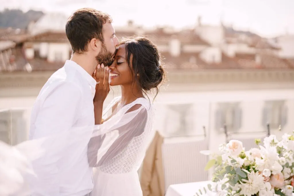 A loving couple embracing and touching foreheads at an outdoor wedding ceremony with floral arrangements in the foreground.