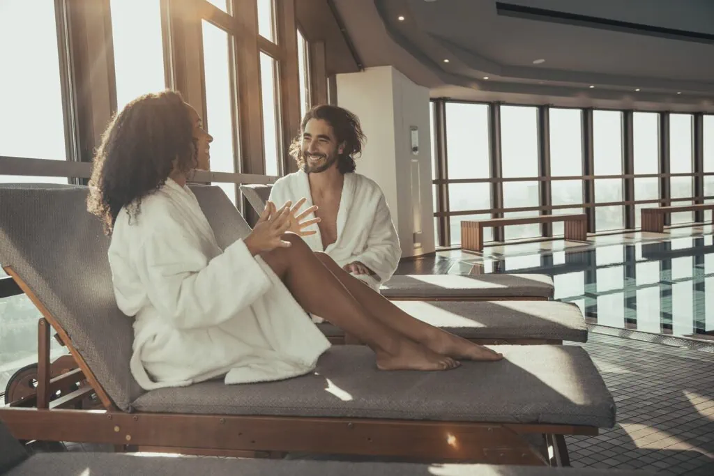Two people in bathrobes engaging in conversation on poolside lounge chairs in a well-lit indoor space.