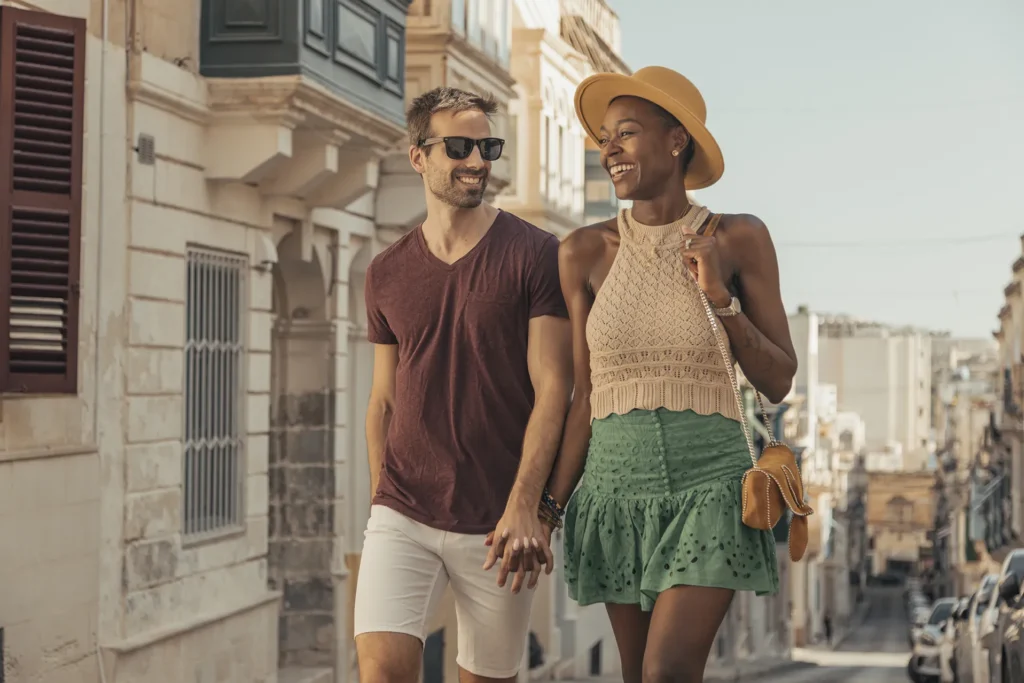 A couple holds hands while walking on a sunny street. The man wears sunglasses, a maroon shirt, and shorts. The woman wears a hat, a beige top, and a green skirt. Buildings are in the background.