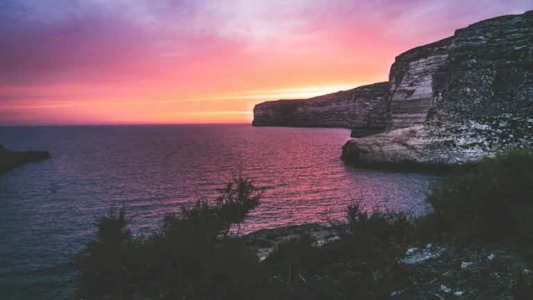 Sunset over a calm sea with dramatic cliffs silhouetted against a vibrant pink and orange sky. Sparse vegetation in the foreground.
