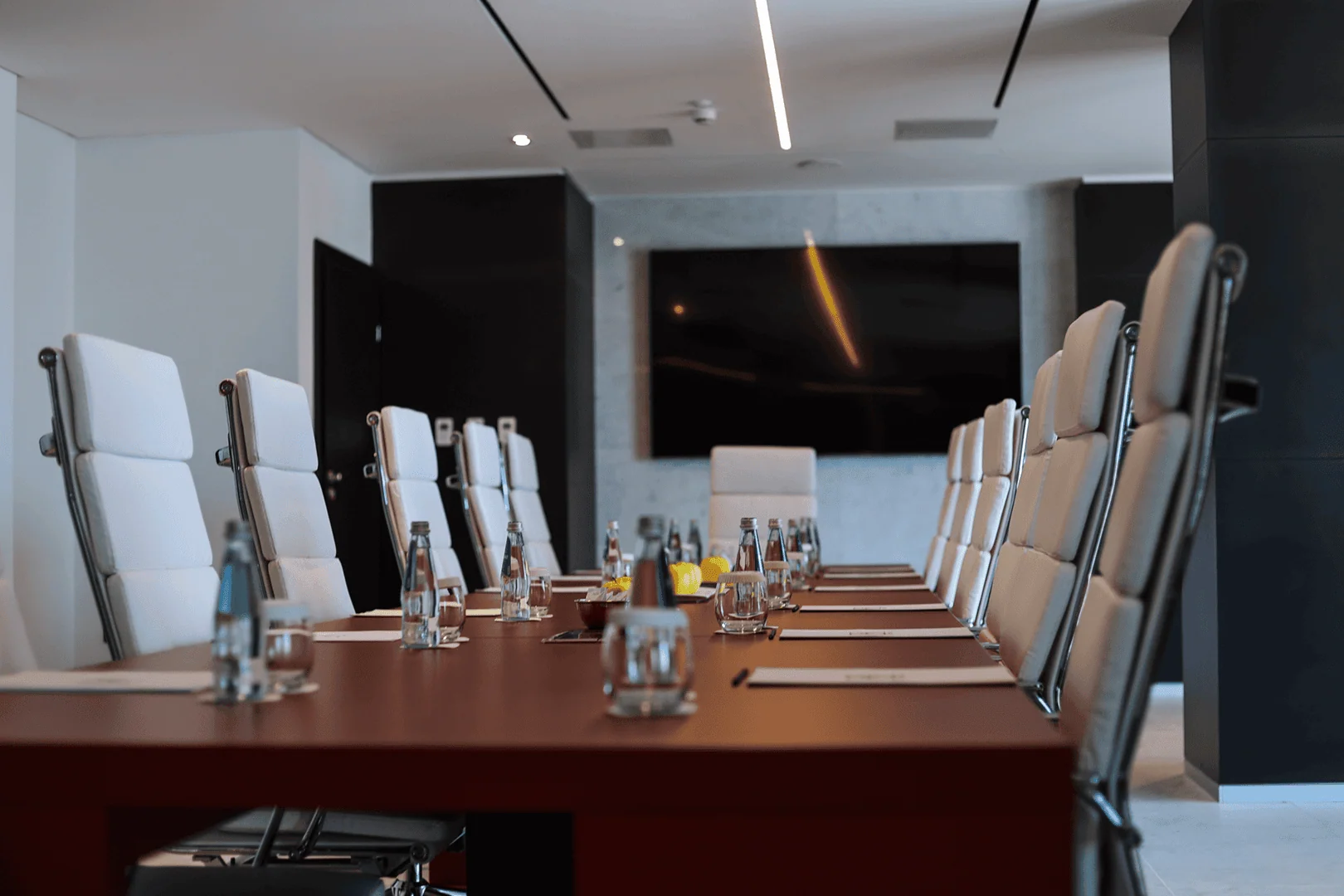 A modern conference room with a long table set with water bottles and glasses, surrounded by white high-backed chairs and facing a large flat-screen TV on the wall.