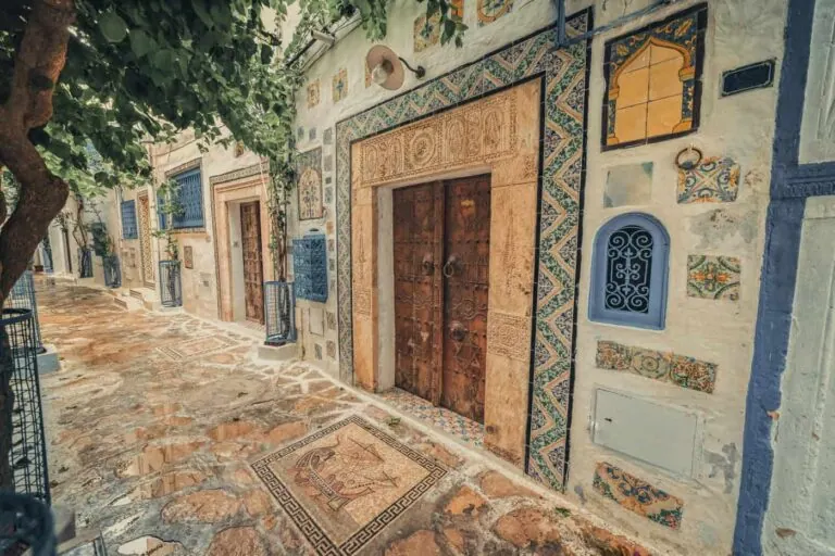 A narrow stone pathway runs alongside an ornate building with mosaic decorations, colorful tiles, and wooden doors, beneath overhanging tree branches.