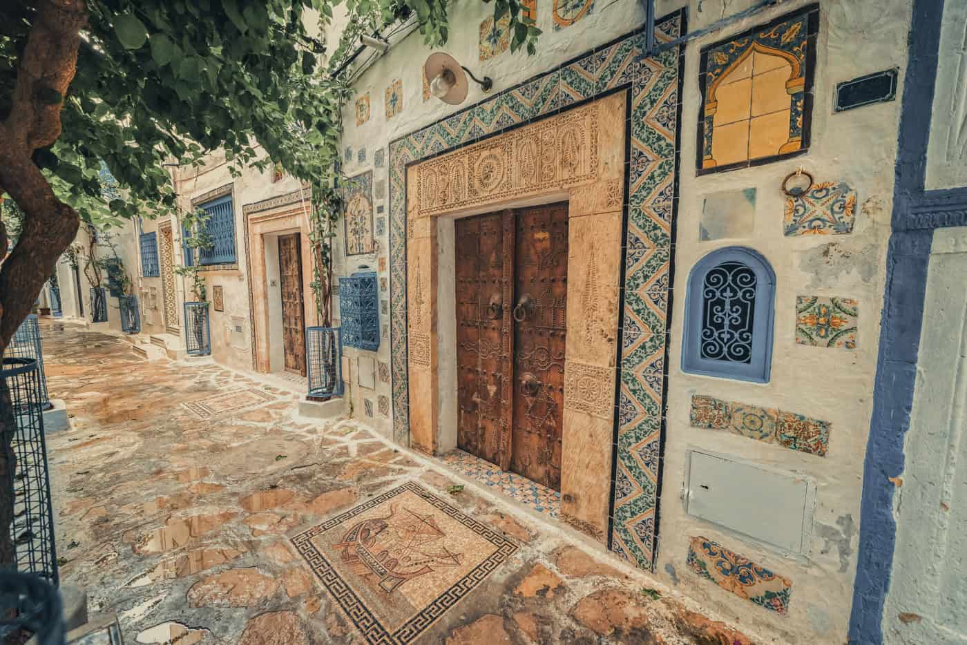 A narrow stone pathway runs alongside an ornate building with mosaic decorations, colorful tiles, and wooden doors, beneath overhanging tree branches.