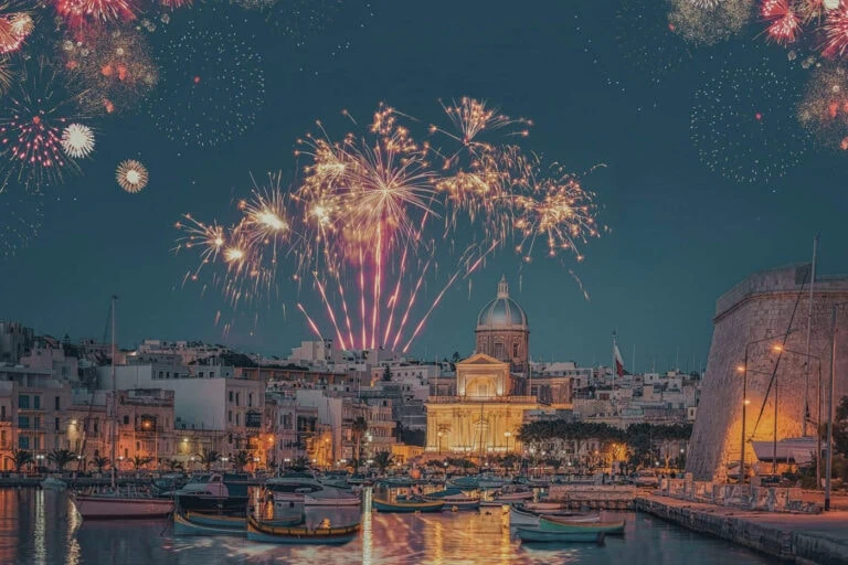 Fireworks light up the night sky over a coastal city with boats docked in the harbor and a dome-topped building prominently in view.