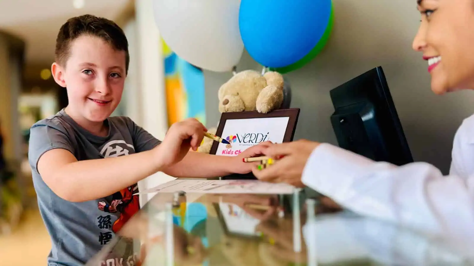 A young boy smiles while receiving crayons from a woman at a reception desk. A sign with "Kid's Club" and balloons are visible.
