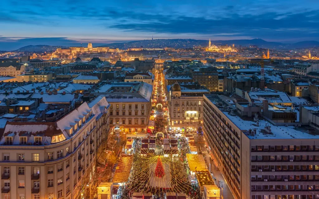 Aerial view of a city at dusk with illuminated streets, buildings, and a central area with festive lights and decorations, surrounded by hills in the background.