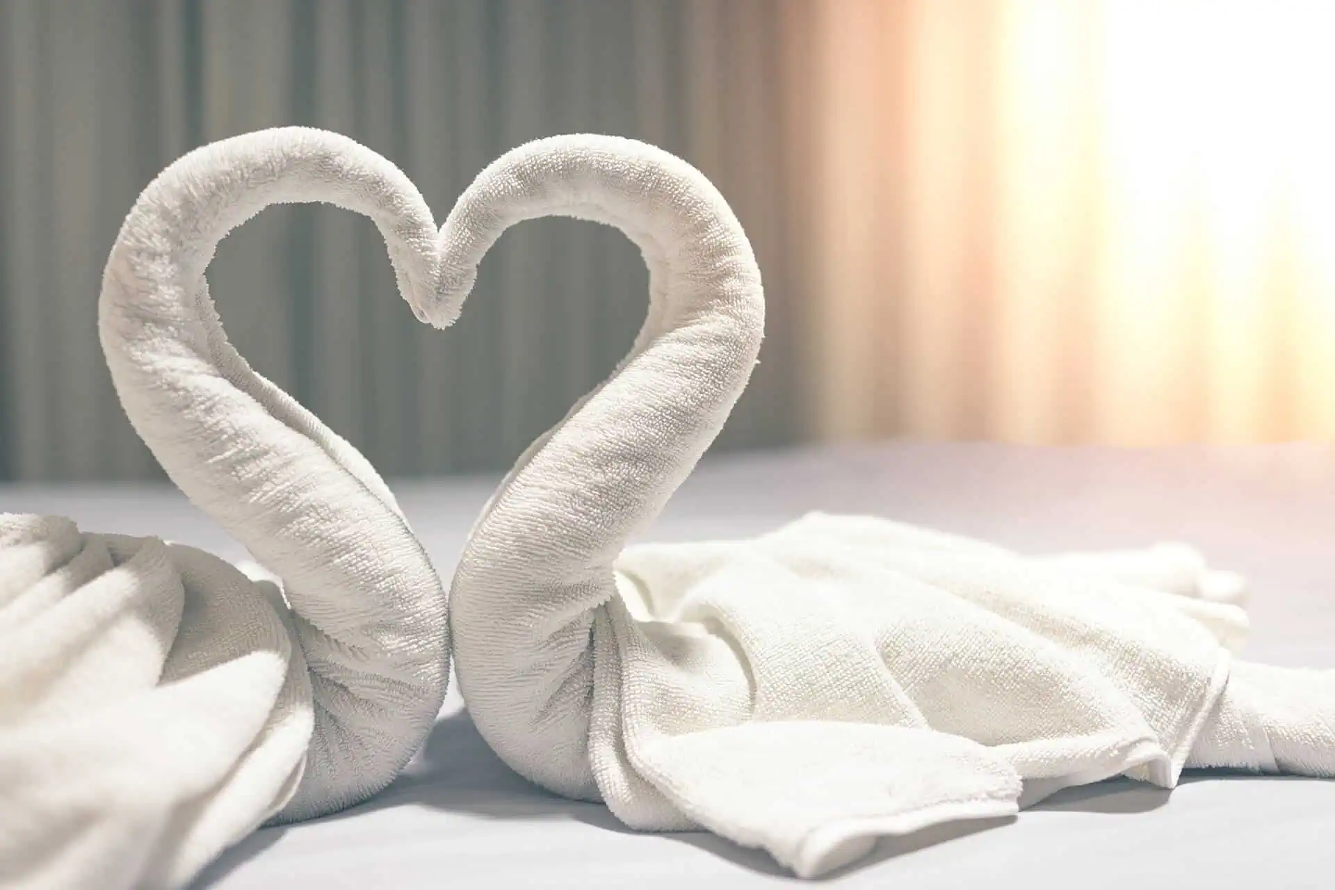 Two towels folded into swan shapes forming a heart on a bed with soft lighting in the background.