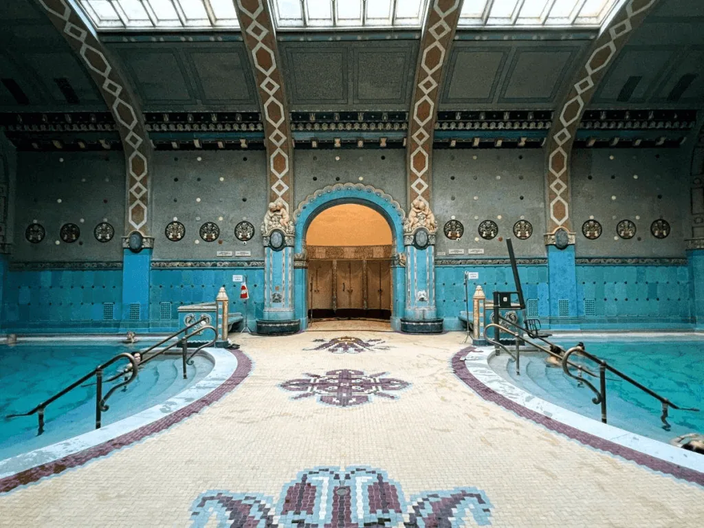 Indoor swimming pool with ornate blue and beige decor, arched entrance, and skylights above. The pool area features mosaic tiles and decorative columns.