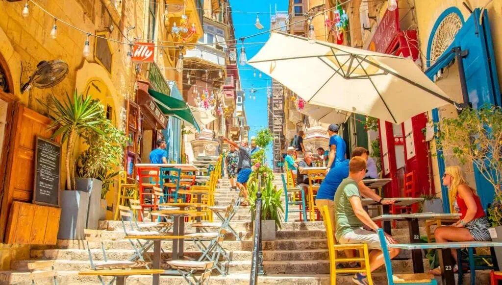 Colorful outdoor cafe on a narrow stone staircase, with people sitting at tables under umbrellas, surrounded by vibrant buildings and plants on a sunny day.