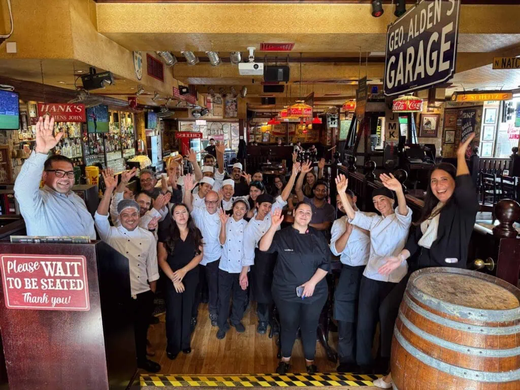 A group of restaurant staff wearing uniforms stand together inside, smiling and raising their hands in a celebratory manner.