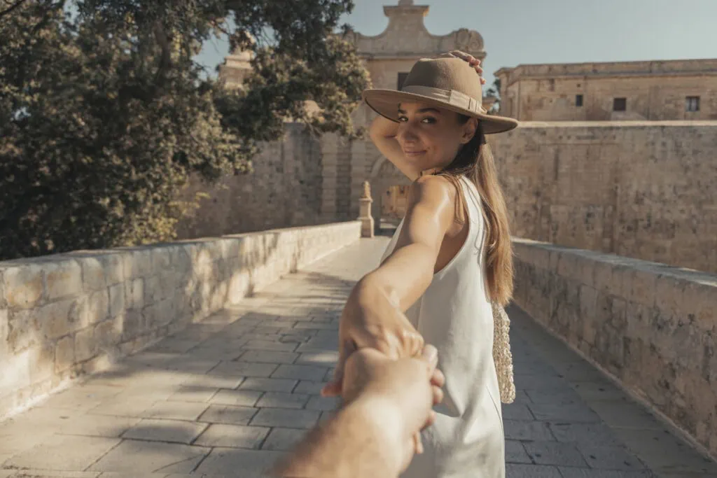 A woman in a hat leads the viewer by hand across a stone bridge, with historic buildings in the background.