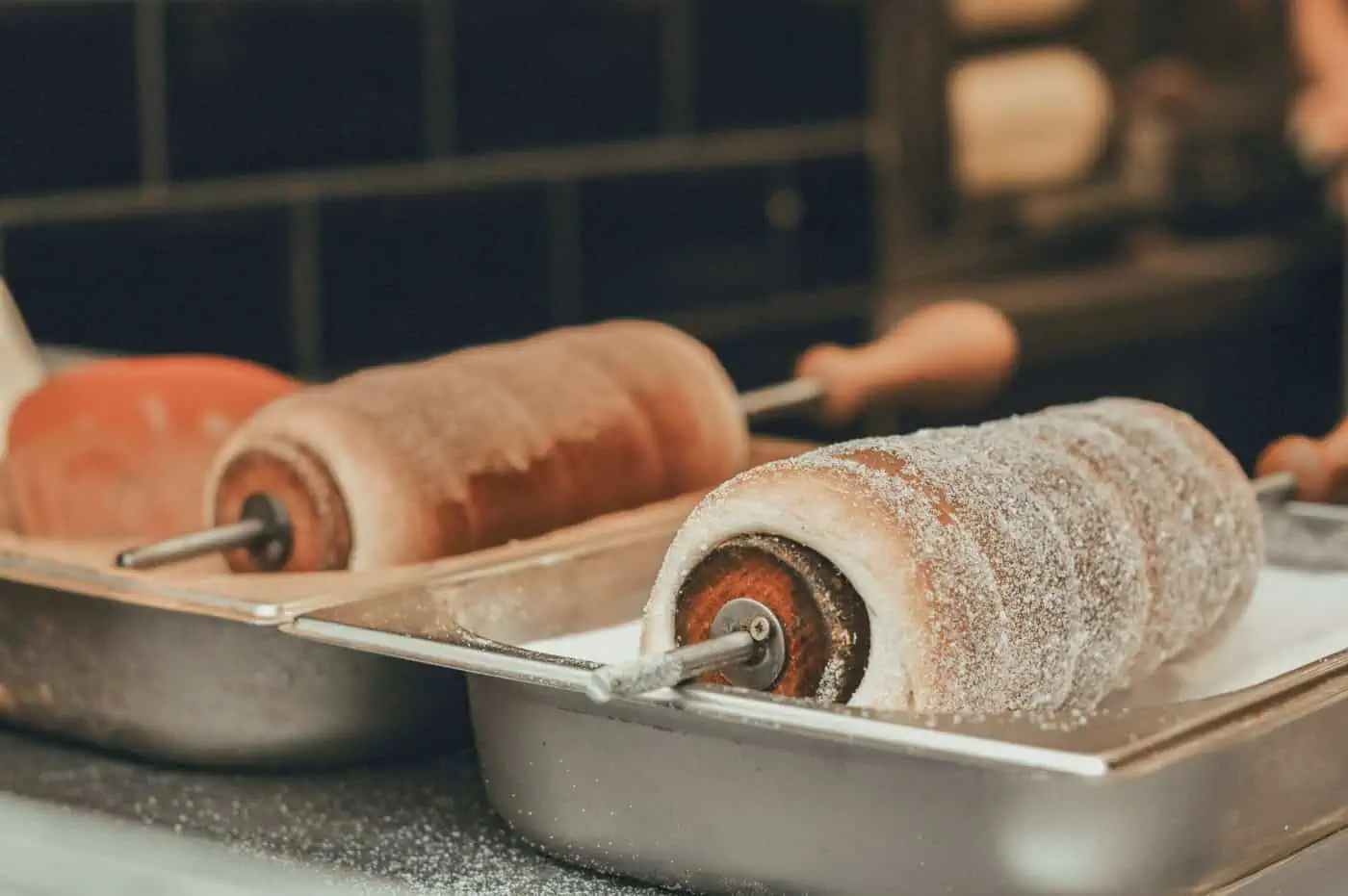 Two kurtoskalacs, or chimney cakes, covered in sugar, are resting on metal trays in a kitchen setting.