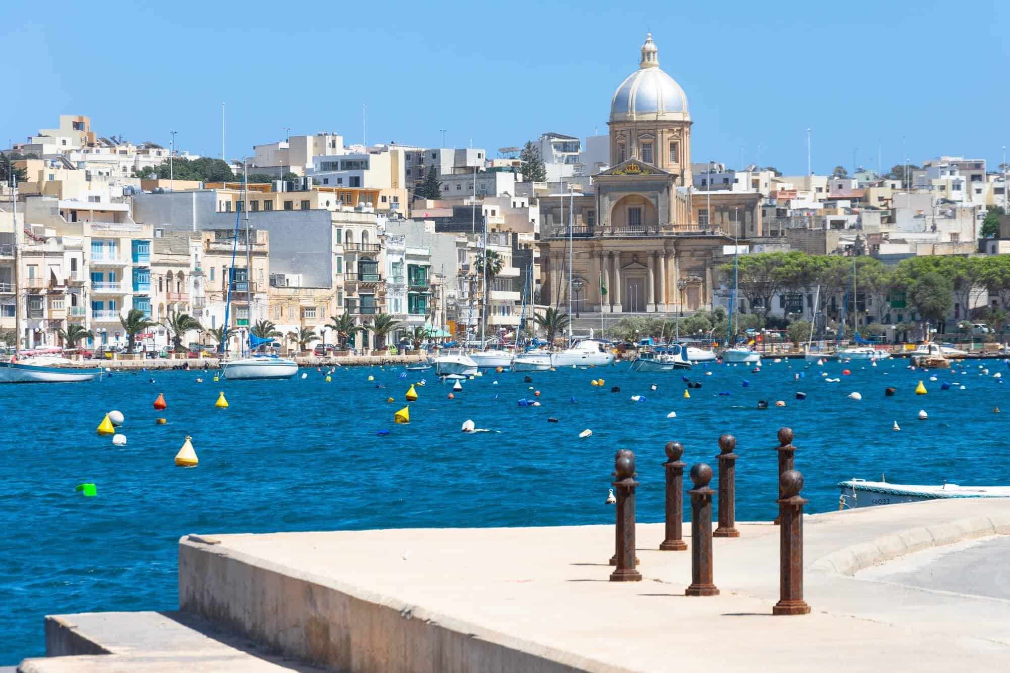 Coastal cityscape with blue water, colorful buoys, and boats. A domed building and multiple structures visible in the background under a clear sky.