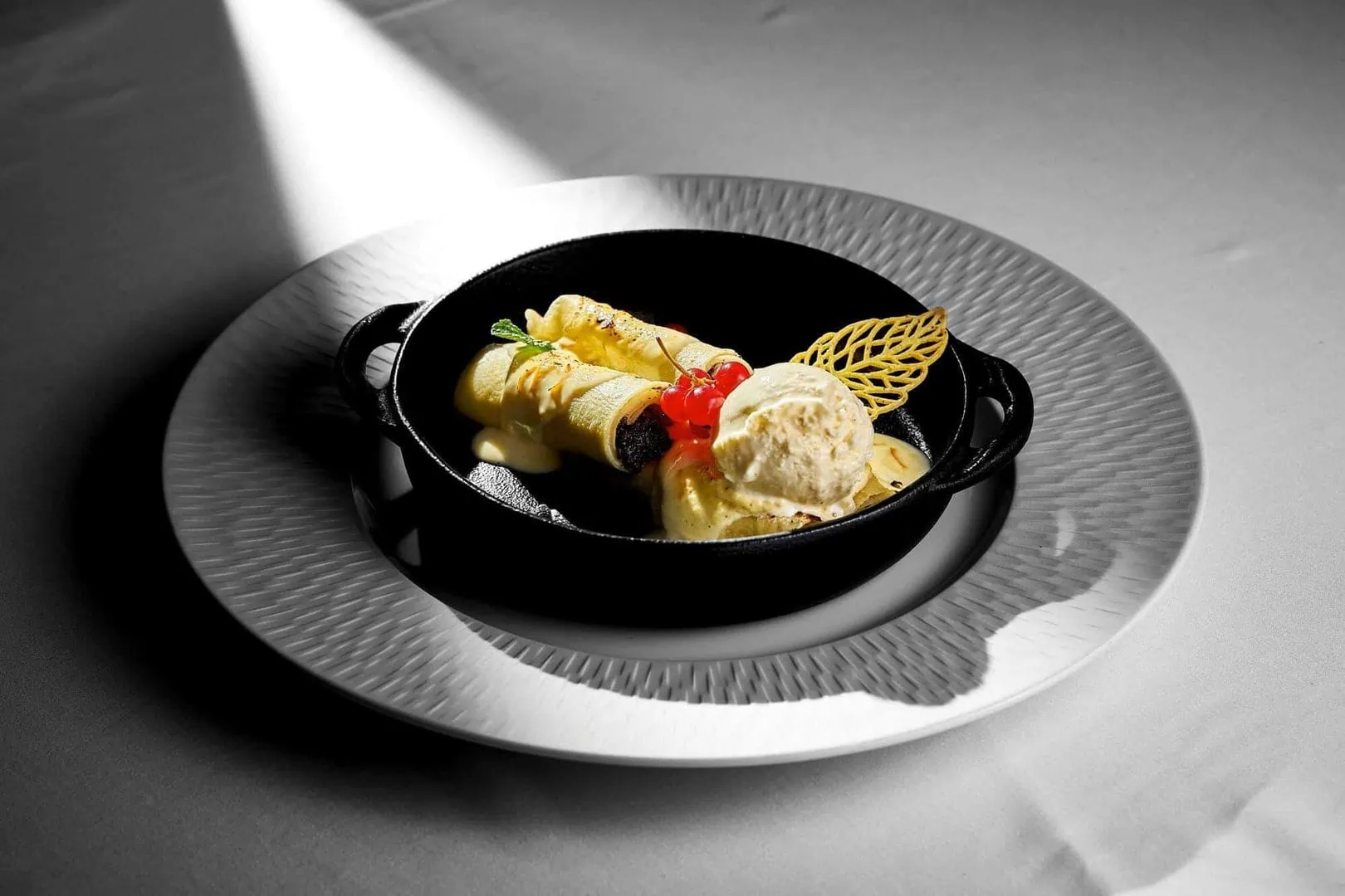 A black skillet on a white plate contains rolled crepes, a scoop of ice cream, red berries, and a decorative wafer. A spotlight highlights the dish against a plain background.