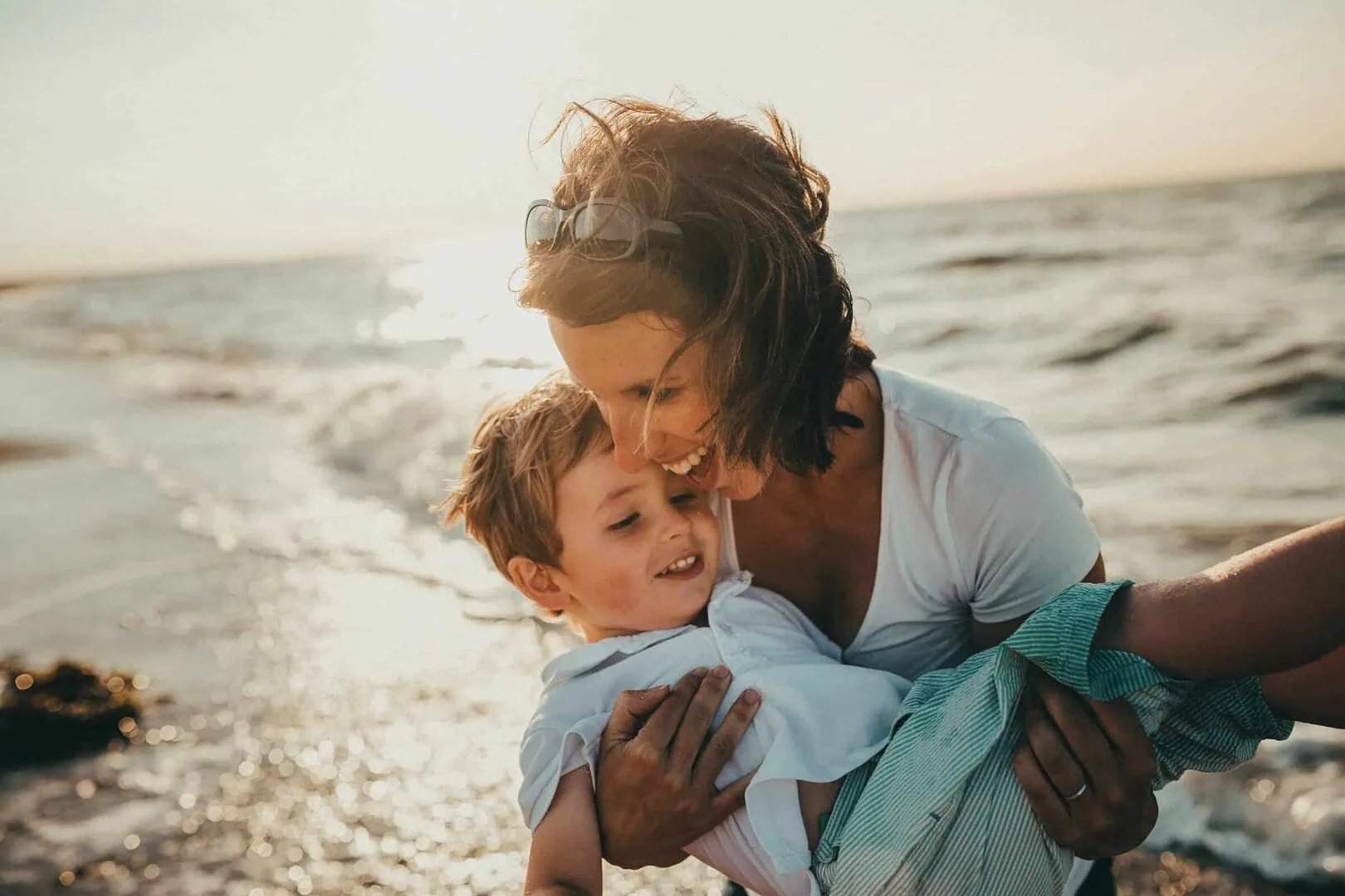 A woman holds a smiling child by the ocean, with waves in the background and the sun setting.