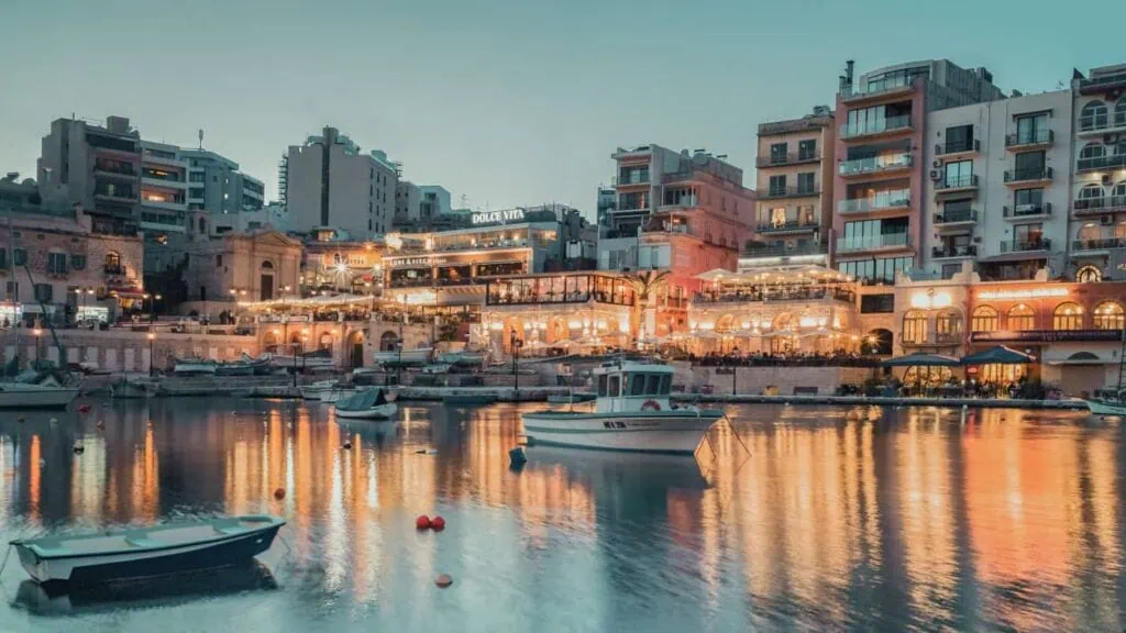Evening view of a harbor with boats in calm water, illuminated buildings, and a clear sky reflecting vibrant lights.