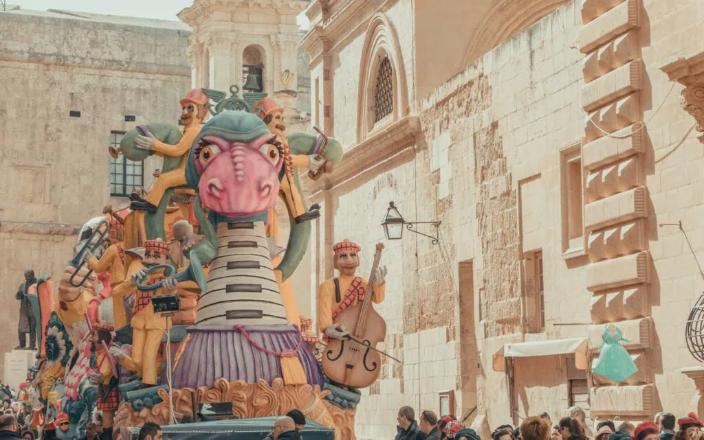 Colorful parade float with large dragon-like figure and musicians in vibrant costumes on a historic street, crowded with spectators.