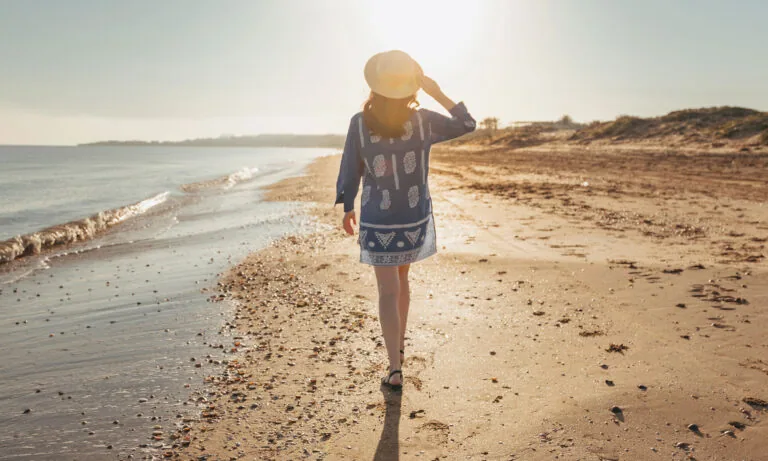 Person in a blue dress and hat walking on a sandy beach toward the sun, with the ocean on the left and a clear sky above.