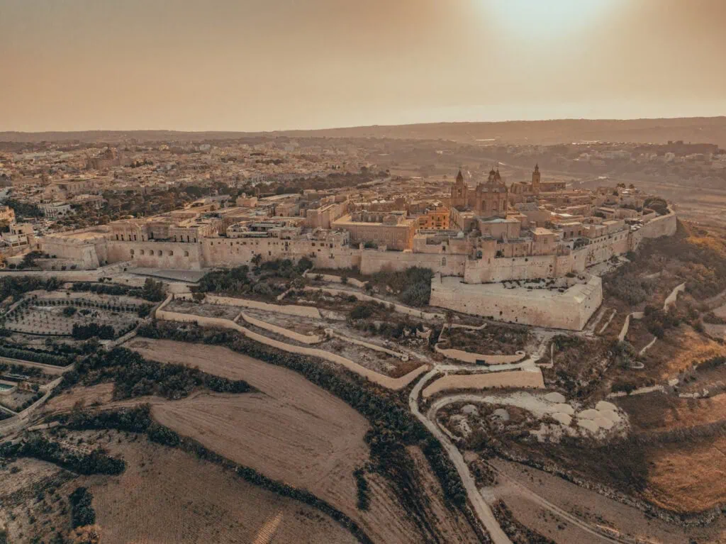 Aerial view of a historic fortified city at sunset, surrounded by fields and modern buildings in the background.