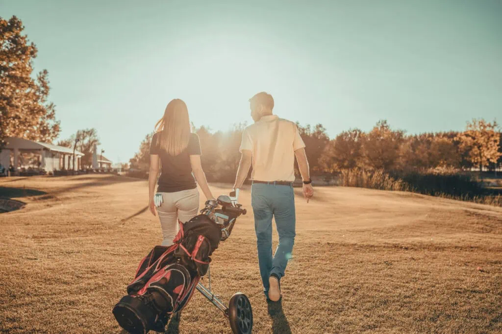 A man and a woman walk across a sunlit golf course, with the man pulling a golf cart.