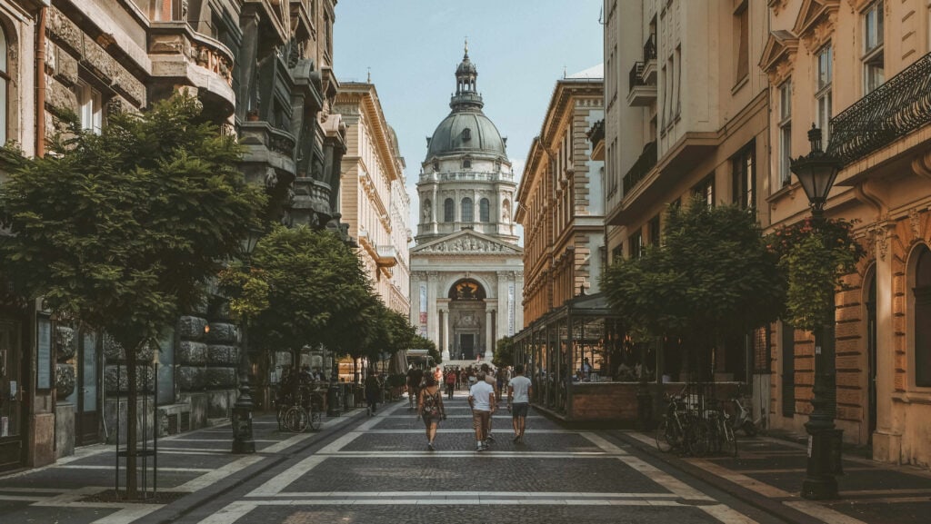 Street view leading to a grand domed building, with people walking along a tree-lined path flanked by historic architecture.