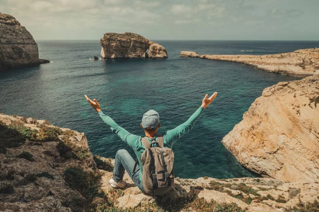 Person sitting on a rocky cliff with arms outstretched, overlooking a blue sea and distant rocky islands.