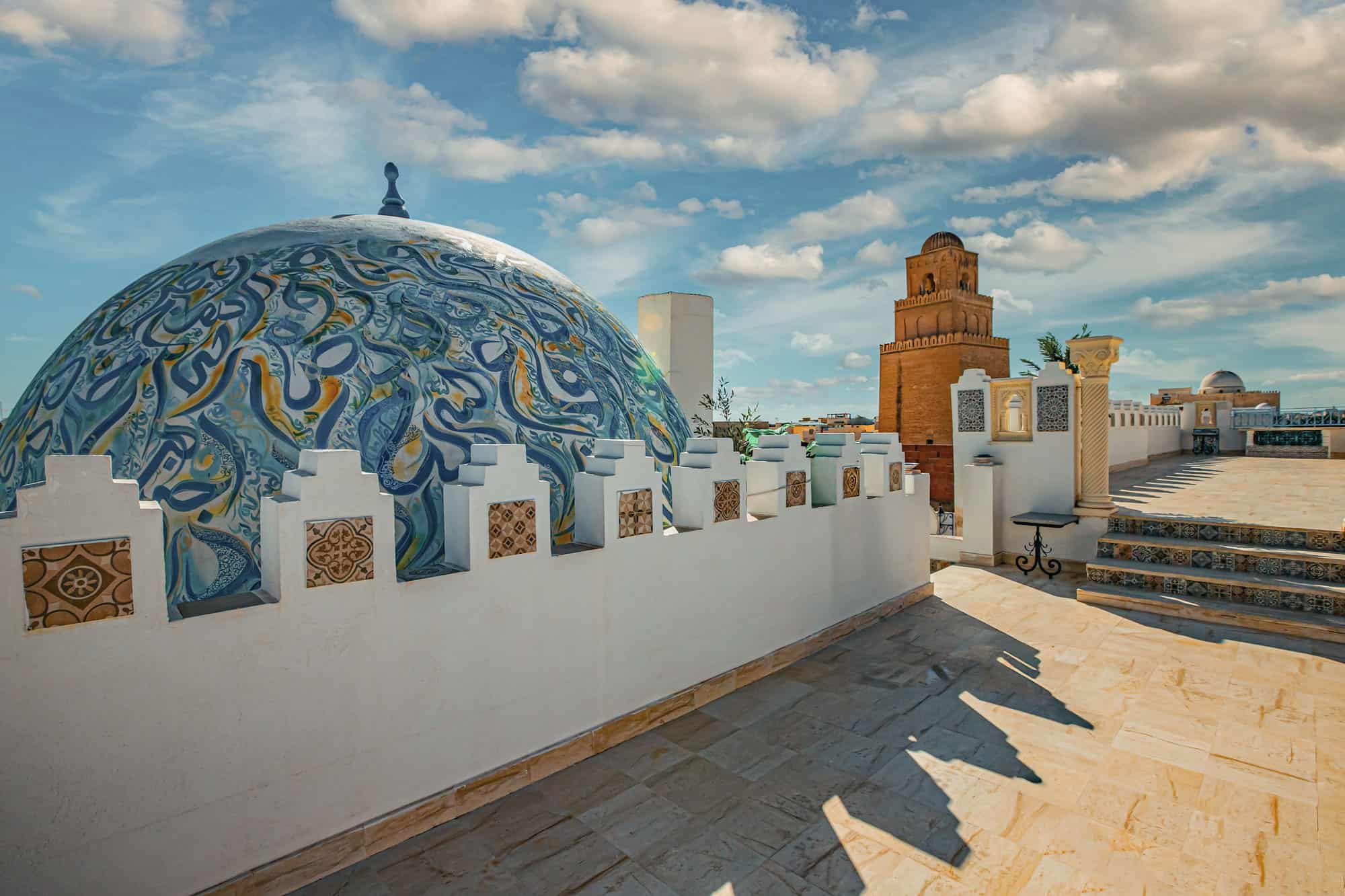 Rooftop view with a decorated dome, white wall, and tower on a sunny day with scattered clouds.