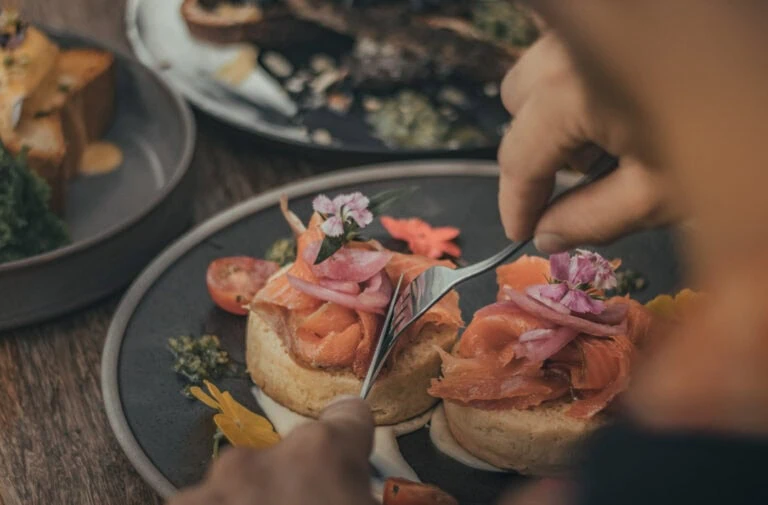 Person using a knife and fork to cut into smoked salmon on a biscuit, garnished with pickled onions and flowers, with other dishes in the background.