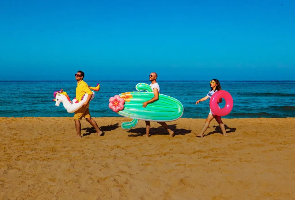 Three people walk in a line on a beach, each carrying a colorful inflatable: a unicorn, cactus, and pink ring. The sky is clear blue, and the ocean is calm in the background.