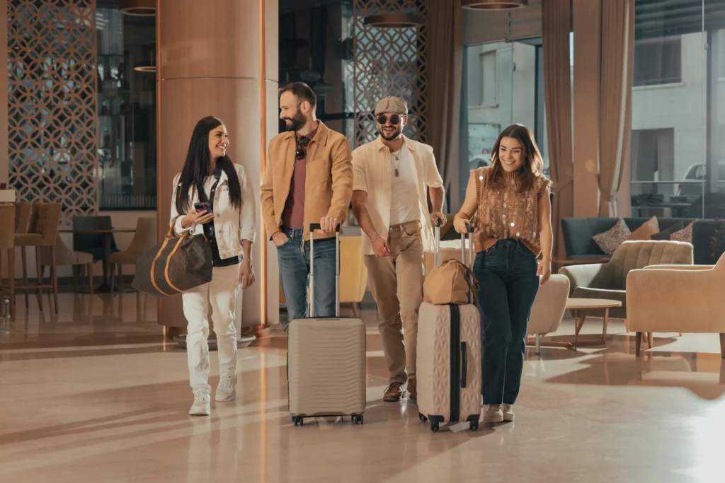 Four adults, two men and two women, walk through a hotel lobby with rolling suitcases and travel bags, appearing to converse and smile.