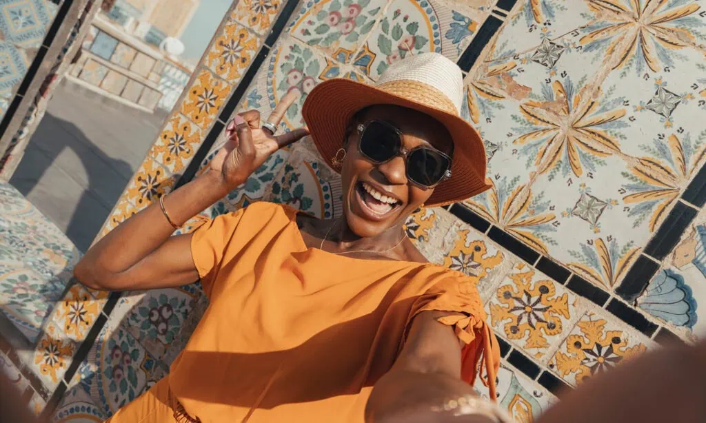 A woman in an orange dress and sunhat smiles and poses for a selfie in front of a wall decorated with colorful, patterned tiles.