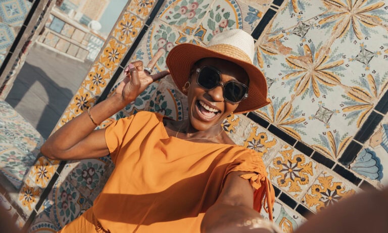 A woman in an orange dress and sunhat smiles and poses for a selfie in front of a wall decorated with colorful, patterned tiles.