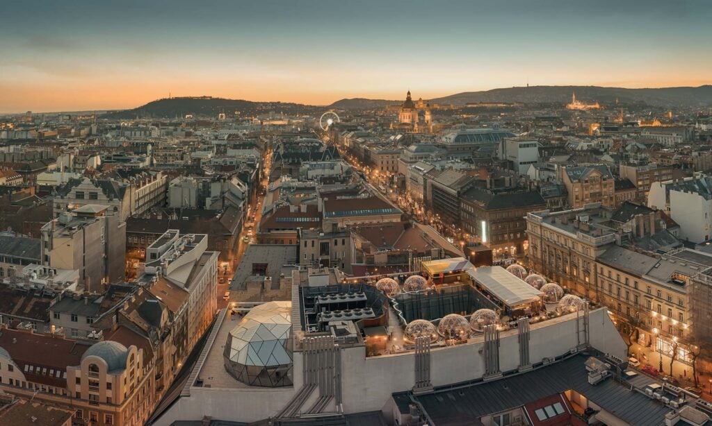 A panoramic view of a cityscape at sunset, with historic buildings, illuminated streets, and rooftop domes, set against distant hills.