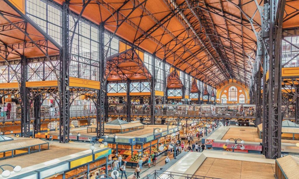 Interior view of a large indoor market hall with high ceilings, iron framework, and multiple vendor stalls on two levels. Shoppers walk among the stalls.