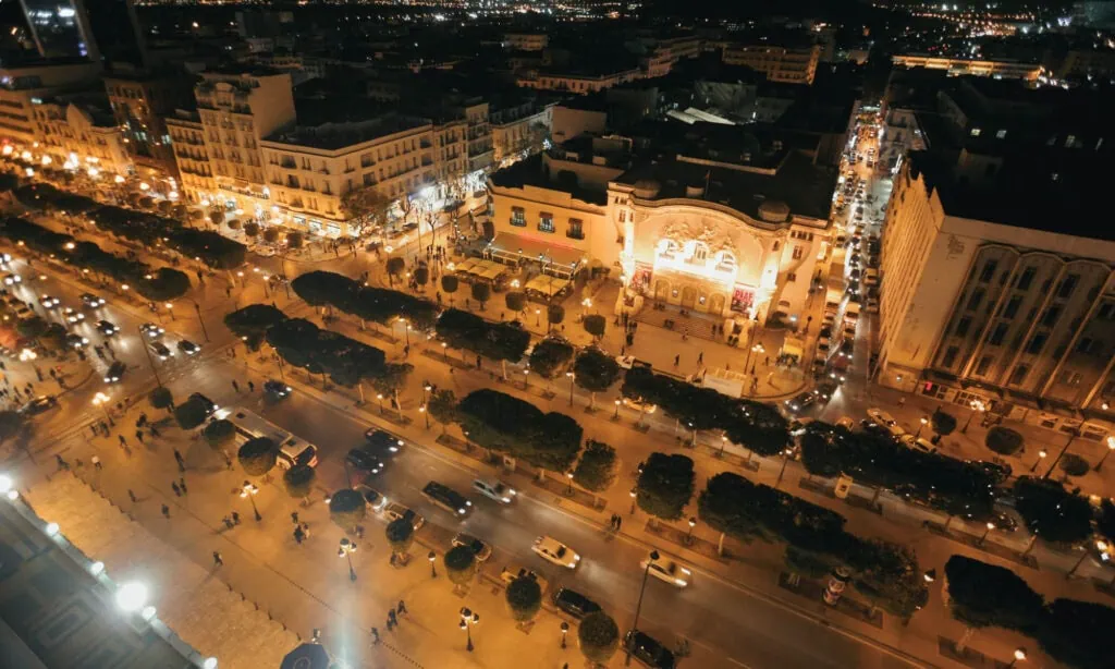 Aerial night view of a busy city street lined with cars, trees, and illuminated buildings, with people walking on sidewalks.
