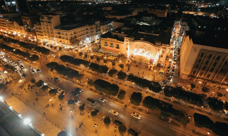 Aerial night view of a busy city street lined with cars, trees, and illuminated buildings, with people walking on sidewalks.