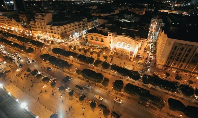 Aerial night view of a busy city street lined with cars, trees, and illuminated buildings, with people walking on sidewalks.