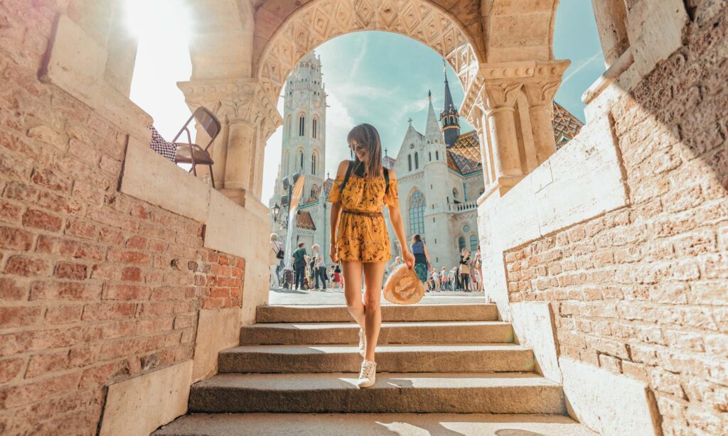 A woman in a yellow dress stands on stone steps under an arched structure, holding a hat, with a church and people in the background on a sunny day.