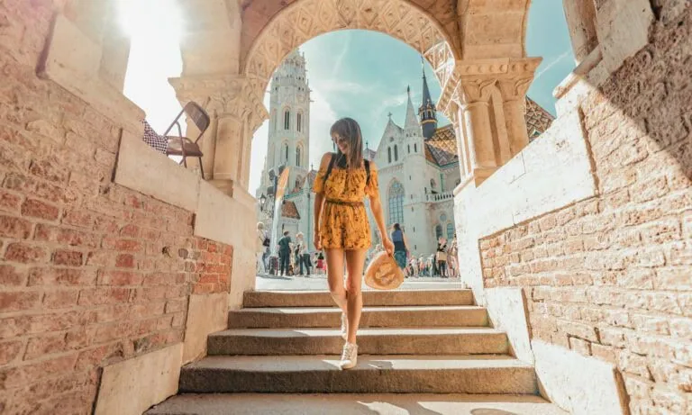 A woman in a yellow dress stands on stone steps under an arched structure, holding a hat, with a church and people in the background on a sunny day.