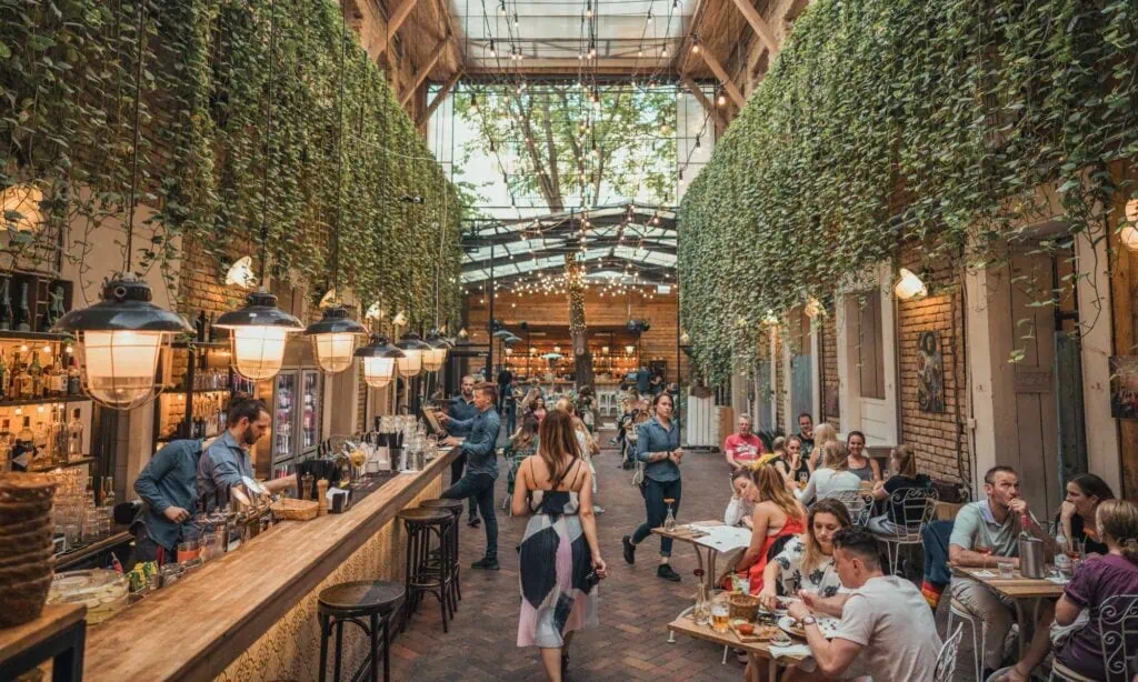 People dine and drink in a lively indoor restaurant with hanging greenery, brick walls, high glass ceiling, and a long wooden bar where bartenders are preparing drinks.