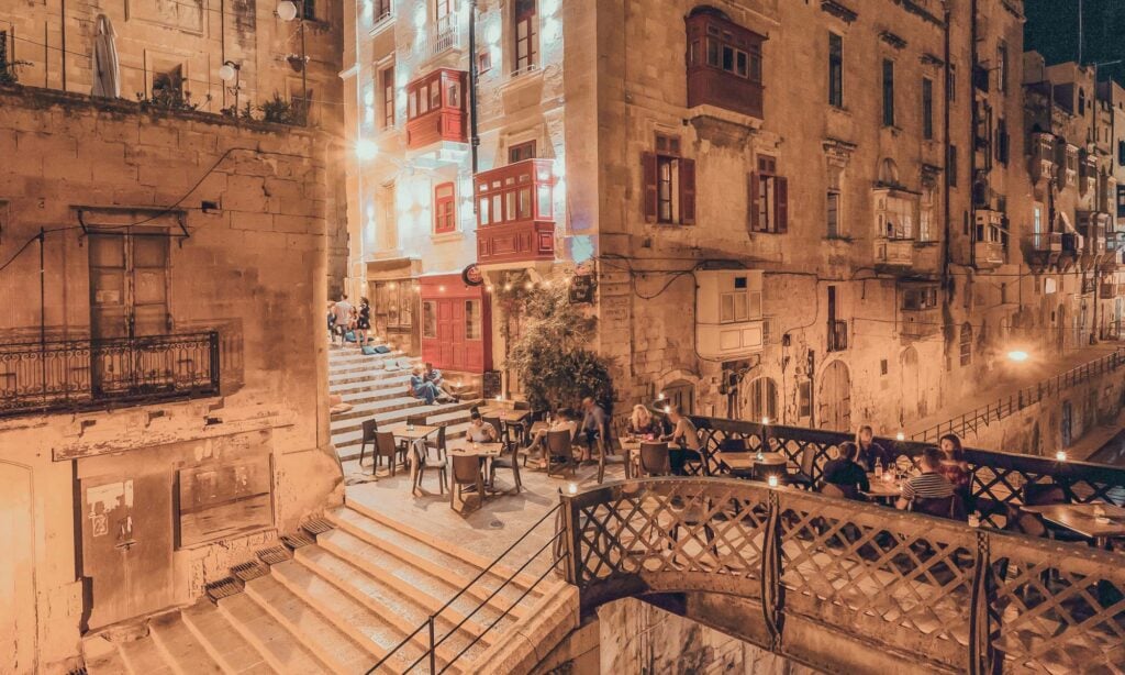 Outdoor tables are set on stone steps and a pedestrian bridge between old, warmly lit buildings at night; people are dining and socializing in the lively urban scene.
