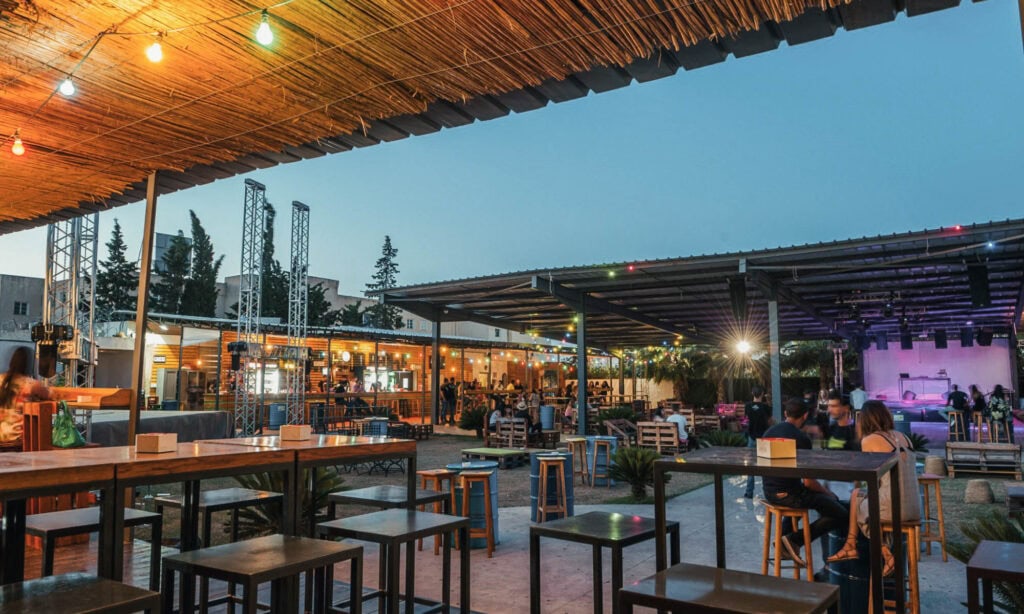 Outdoor bar and dining area with string lights, wooden tables and stools, people sitting, and a stage in the background at dusk.