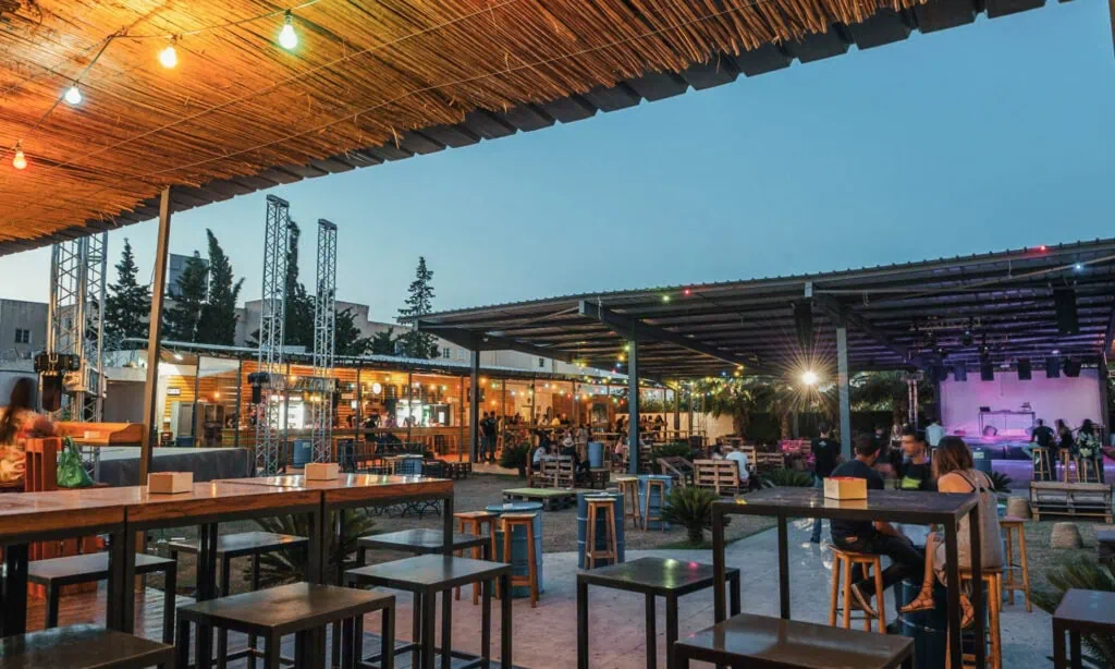 Outdoor bar and dining area with string lights, wooden tables and stools, people sitting, and a stage in the background at dusk.