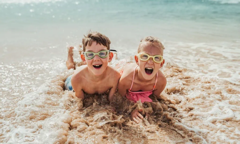 Two children wearing goggles lie on wet sand at the beach, smiling and laughing as ocean waves wash over them.